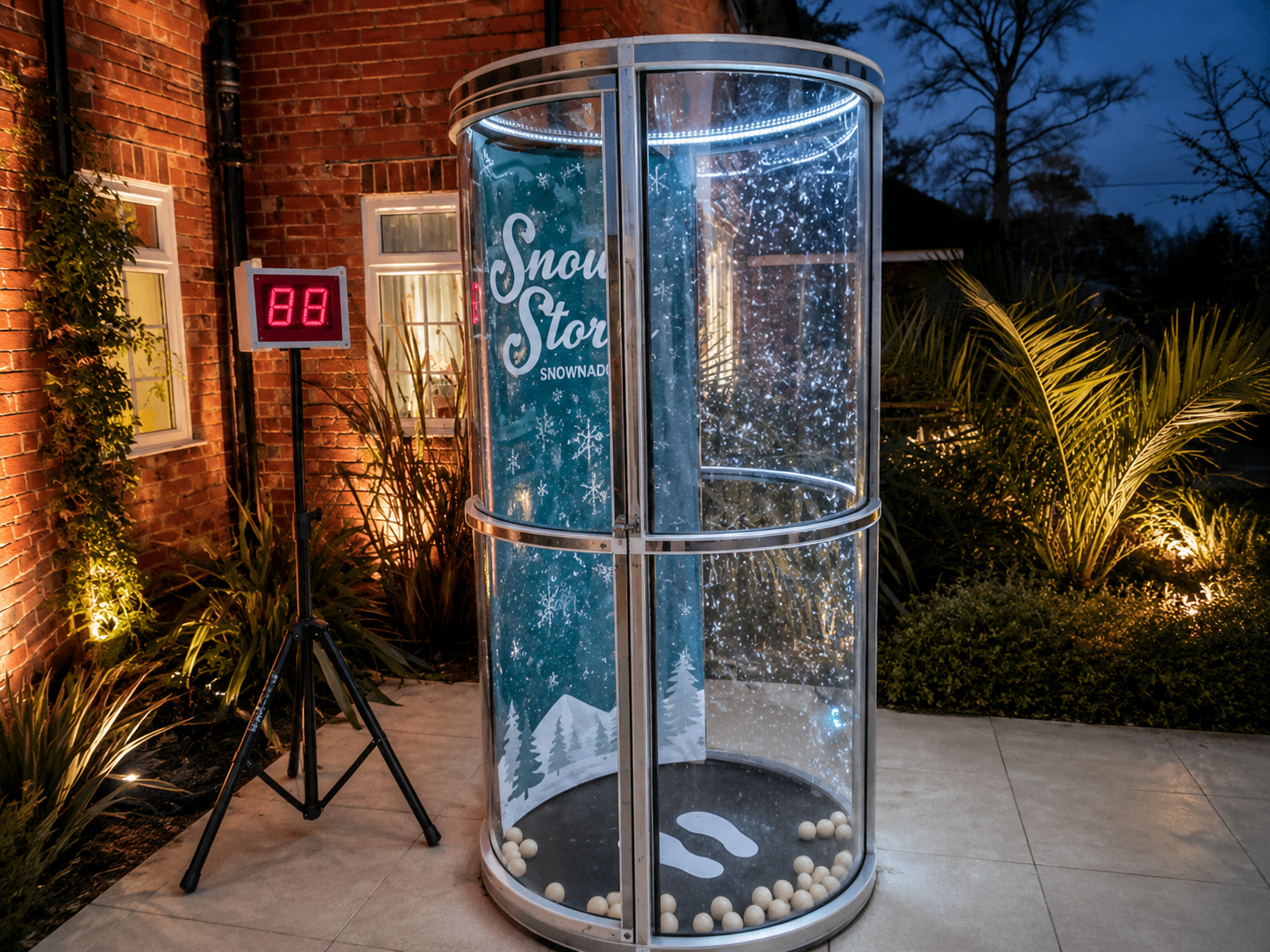 Winter-themed cylindrical glass display labeled 'Snow Store', with falling snowflakes and white balls at the base, next to a brick building; a tripod with a red LED '88' display nearby.