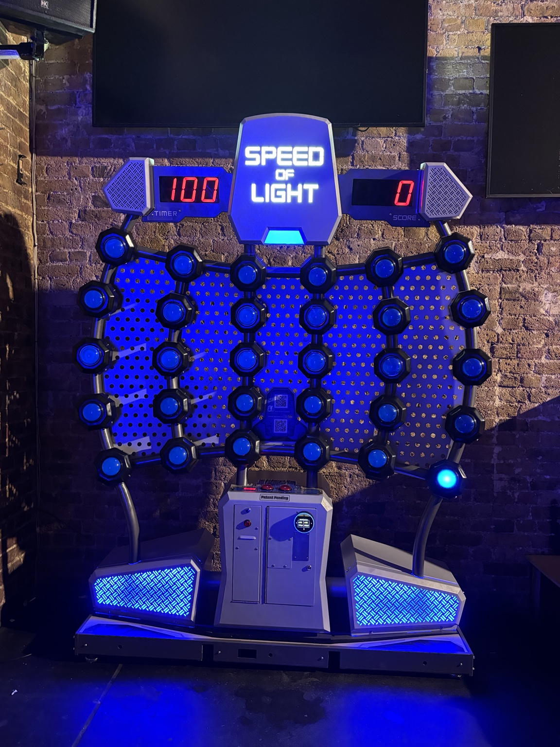 Speed of Light arcade game: a blue-lit hexagonal light grid with a centralSpeed of Light panel against a brick wall, timer readout on the left shows 100 and score on the right shows 0.