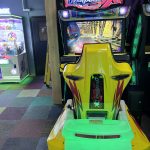 Bright yellow-green arcade racing game cabinet with a large screen showing a city road, and a neon 'Overtake X' sign above the cockpit.