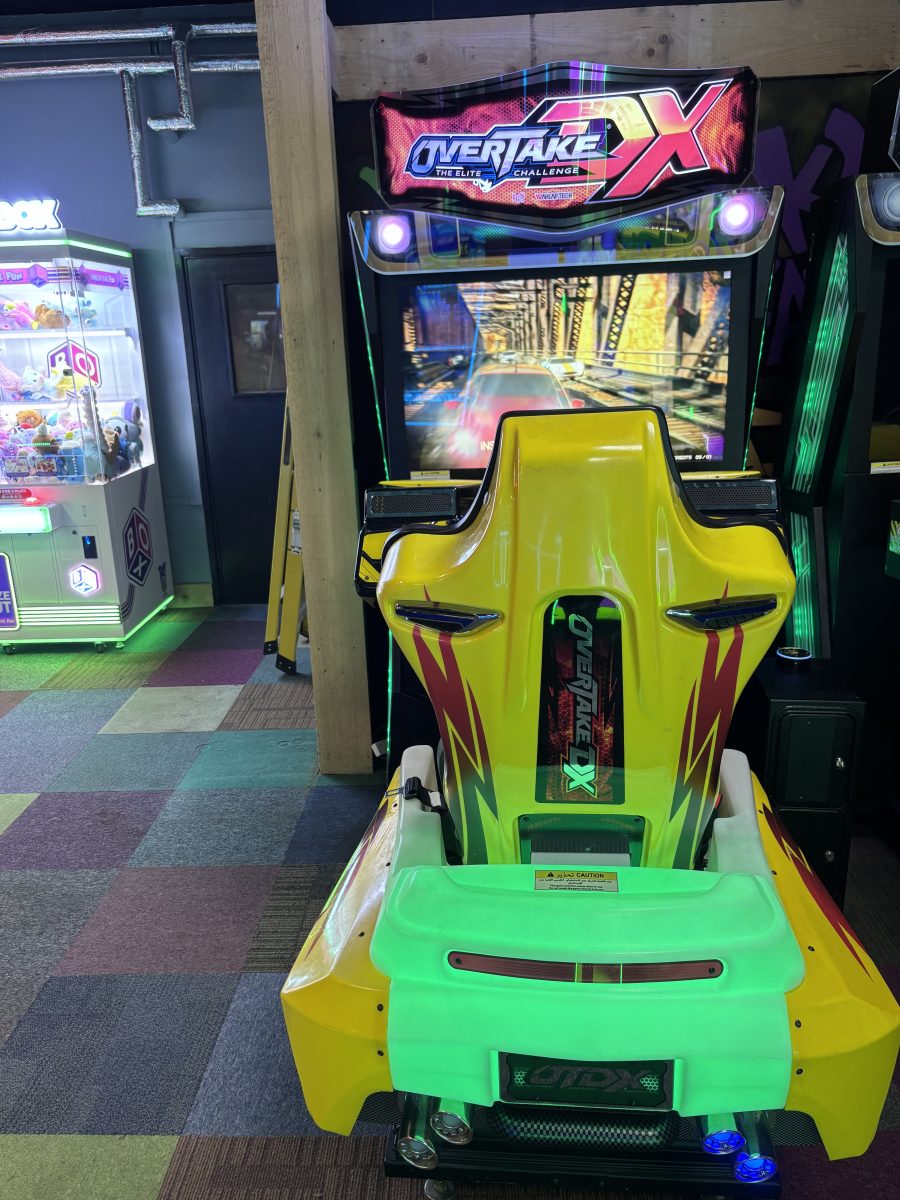Bright yellow-green arcade racing game cabinet with a large screen showing a city road, and a neon 'Overtake X' sign above the cockpit.