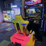 Yellow and pink arcade car race simulator in a neon-lit game room with other claw machines in the background intent on a driving game setting