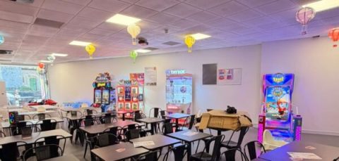 Colorful arcade classroom with rows of black tables and chairs, vending machines and arcade games along white walls, and hanging lanterns from the ceiling.